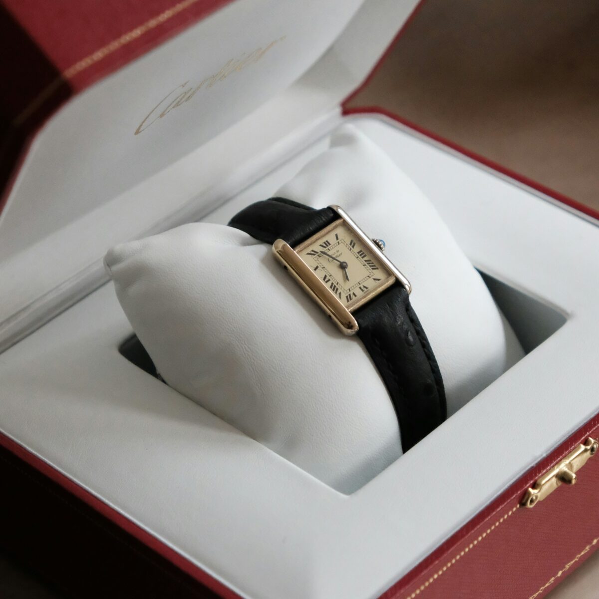 Must de Cartier Tank with a gold-tone rectangular case, Roman numeral dial and black leather strap in an open red presentation box.