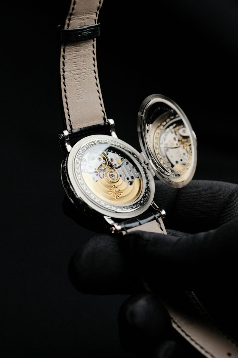 Patek Philippe watch with its hinged caseback open, revealing the movement and gold-tone rotor above a black leather strap.