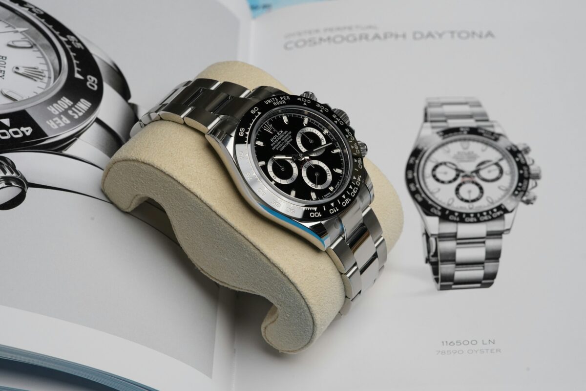 Rolex Daytona with a black dial, silver subdial rings, black tachymeter bezel and steel bracelet on a cream cushion over a catalogue.
